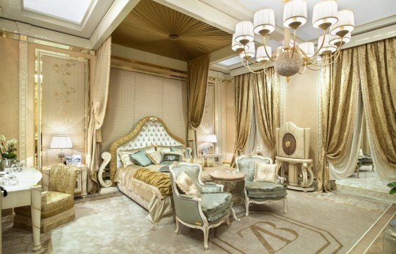 This picture shows a modern and luxurious bedroom designed with an open concept. It features a four poster bed upholstered in blush pink velvet, a tufted headboard, plush white bedding, and gold accents. There is an ornate, mirrored wardrobe to the left, and a set of French doors leading to a private balcony. The walls are painted a soft white, and the floor has a light-colored hardwood finish. A round glass table with a statement lamp sits in front of the bed, and there is a chaise lounge at the foot of the bed.