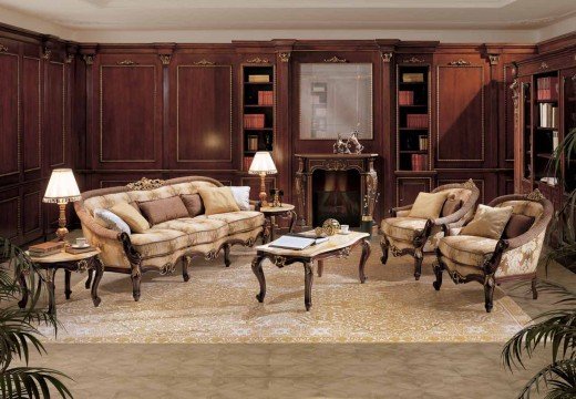 This picture shows an opulent living room with a luxurious velvet sofa, a tufted armchair, and a crystal chandelier. The walls are painted in a soft muted grey color, and the floor is lined with dark hardwood. The room also features mixed metal accents that add to the overall sophistication and glamour of the space.