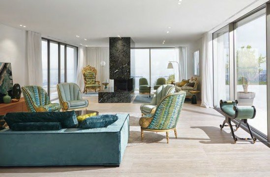 This picture shows a luxurious interior design of an open-concept living room. It features a large, curved beige sofa and armchair set with a glass coffee table and an area rug in the center. There is also a stylish modern fireplace in the corner with a wooden mantelpiece, and two large windows on either side of the room with white drapery and sheer curtains. The walls are decorated with framed artworks, and accent lighting fixtures are seen throughout.