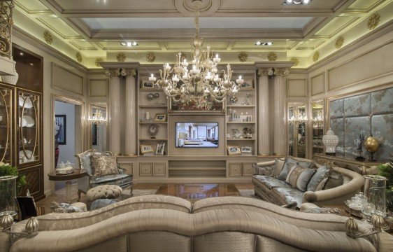 This picture shows an elegantly designed, modernly decorated living room. The room features a large cream-colored couch with several decorative pillows, a glass coffee table, and a patterned rug. There is also a large bay window with sheer white curtains covering it, as well as several wall hangings near the windows. The walls are painted a mix of neutral colors, and the room is lit by several modern chandeliers.