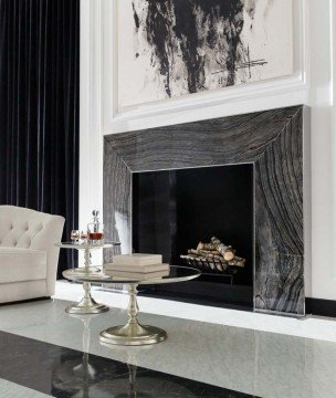 This picture shows a luxurious, modern living space with a grand double-sided staircase framed by two white marble-clad columns. The staircase leads down to an open plan living area with a tiled floor, beige sofas, and glass coffee tables. A large mirrored wall with a geometric shape reflects the furniture and leads the eye out towards a tall arched window framed with curtains.