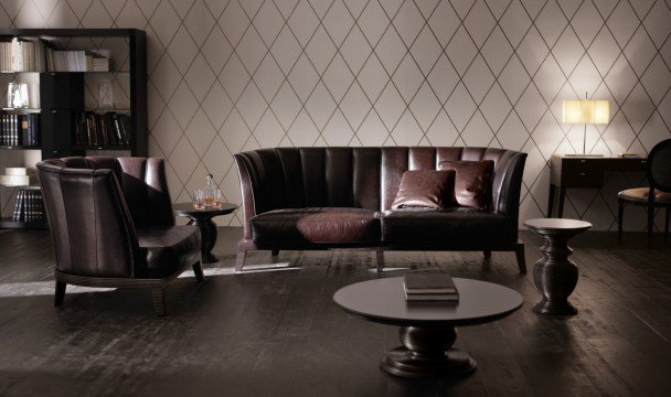 This picture shows a luxurious bedroom interior. It features a giant bed with a light grey headboard, a matching footboard, and white bedding. The walls are decorated in a dark grey, and the floor is covered with a light grey rug. On either side of the bed are two tall nightstands, with large table lamps and decorative glass dishes. At the foot of the bed is an upholstered bench, and on the wall behind it is a large framed mirror.