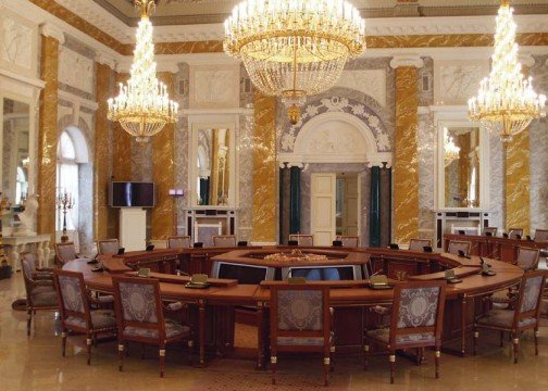 This picture shows an interior design in a large, luxury bedroom. The bedroom has light cream walls and ornate crown moulding along the top. The room is filled with pieces of classic furniture such as a four-poster bed covered in a white bedspread, two chairs with footstools in the corner, and a tall armoire with intricate carvings near the entrance. On the side of the room is a luxurious chaise lounge chair with plush green upholstery for lounging. There are also two traditional side tables with lamps and fresh flowers in vases placed on