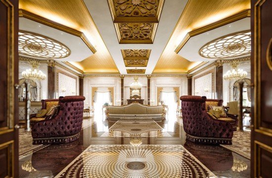 This picture shows a luxurious living room interior with rich gold and maroon accents. The room has a modern and elegant design, with a large brown velvet couch against the wall, accented by two matching armchairs; a round marble topped side table in between them for extra seating. The walls are beige with a light-colored pattern that adds an air of sophistication. There is a luxurious maroon accent rug in the center of the space, and a grand chandelier hanging from the ceiling. The artwork on the walls is striking and adds to the overall opulence of the room.