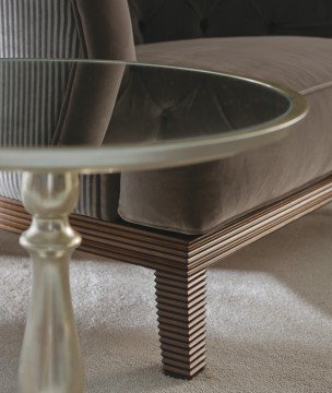 This picture shows the inside of a modern and luxurious living room. The room is decorated with a white and beige color scheme and accentuated by gold and black accents. There are comfortable beige sofas, two white armchairs, and a marble-topped coffee table with a glass top in the center. A large, unique golden chandelier hangs from the ceiling, and a black and gold area rug lies on the floor. The walls are lined with golden wall hangings and a large mirror above the sofa adds to the regal decor.