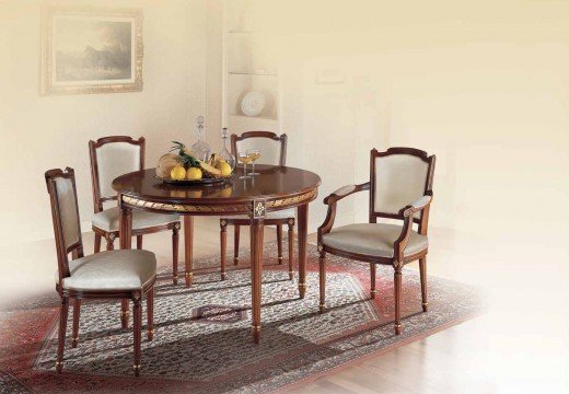 This picture is of an elegant formal living room with a modern design. It features a white sofa with two side chairs and a single armchair in the front, all upholstered in a light gray fabric. The focal point is a large wall mural in beige and gold tones. A gold coffee table with glass top stands in the middle of the seating area and a modern glass chandelier hangs above it. The walls are painted white and feature several other gold accents.