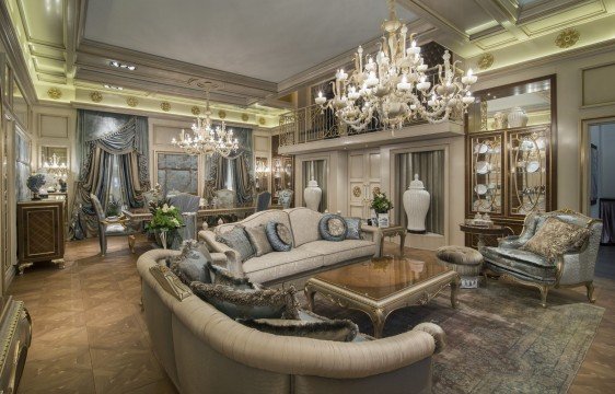 This is a picture of a modern luxury bedroom in neutral colors. The room features beige walls, a large bed with ornate headboard and white bedding, a nightstand, a chaise lounge chair, a window with cream curtains and sheer drapes, and an area rug in shades of gray and beige. In the corner there is a tufted upholstered armchair with a white fur throw blanket and a side table with a vase of flowers.