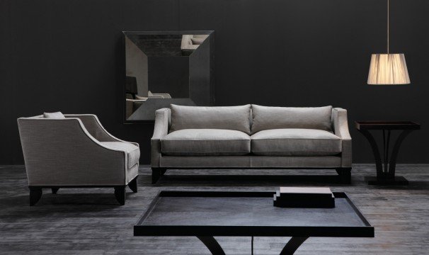 This picture shows an elegant living room with a rich color palette of gold and white. The floor is marble, with a large white rug that defines the sitting area. Two beige sofas are situated around a white and gold coffee table in the center of the room. On the left side is a large gold wall mirror and two white armchairs. On the right side is a white console with two chairs and a gold and white abstract painting above them. The room is finished off with two green plants.