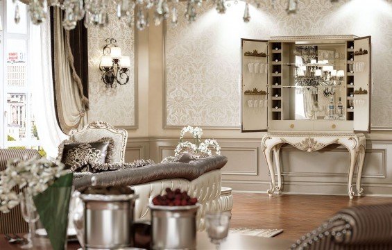 This picture is of an ornately decorated modern bathroom. The room features a luxurious marble countertop with oval sink and gold faucet, marble flooring, a large jacuzzi bathtub, glass shower area, a chandelier, and a dark wooden vanity with several drawers and a shelf. There are also two padded benches upholstered in ivory fabric.