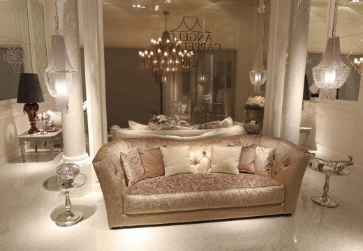 This picture shows a luxurious and modern living room in a home. It features a large, light grey sectional couch with purple and white pillows, a contemporary blue chandelier, a stunning marble accent wall, and an elegant glass and gold coffee table. The interior design is further enhanced by the addition of a variety of stylish gold and black furniture pieces and accessories, as well as various framed artwork and decorative plants. The overall look is one of sophisticated elegance and opulence.