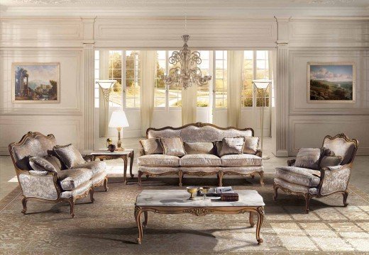 This picture shows a modern luxury living room. The room has light-colored marble floors, and white walls adorned with hanging framed art pieces. On the right side is a gray sofa with two armchairs in a bright red fabric. In the center of the room is a round glass coffee table surrounded by a circle of white chairs. The back wall features a wall-mounted television and shelves filled with books and decorations. On the left side is a large window with thick beige curtains. The overall look of the room is bright, airy, and modern.