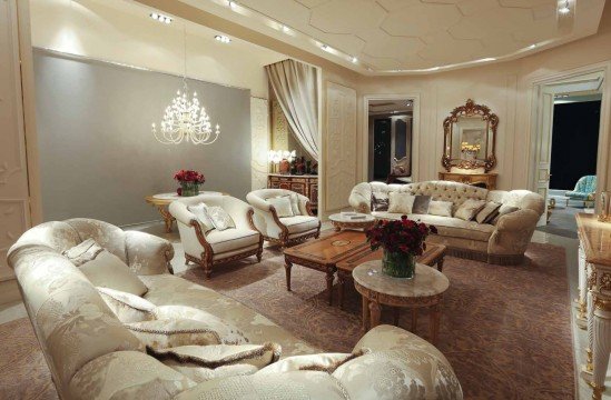 This picture shows a luxurious bedroom with beautiful decor. The walls are painted in a light beige color, and the floor is covered in a white and grey patterned rug. The bed has a cream colored headboard and footboard and is topped with a white tufted comforter. A large window is on one wall and is framed in dark wooden panels. On the opposite wall is a metal framed mirror and an ivory colored armchair. In the corner of the room stands a tall plant in a wooden urn.