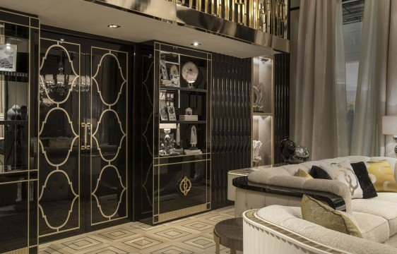 This picture shows a modern, luxurious living room. In the center of the room is a large, round chandelier with intricate details. There are two deep brown leather couches flanking either side of the chandelier and a glass coffee table in between them. The walls are lined with a cream-colored wallpaper that has gold accents. On one wall there is a television mounted on the wall with a decorative wall sconce next to it. The floor is covered in a plush, white rug and there is a large window with sheer curtains on the far wall.