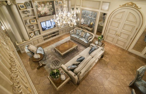 This picture shows a luxurious living room with high ceilings, exquisite furniture, and a grand chandelier. The walls are covered in light gray velvet and the floors are covered in a white and gray patterned rug. There are several pieces of furniture including two armchairs, an ottoman, and a white sofa surrounded by several ornate pillows. The walls are adorned with golden frames containing artwork and the center of the room is illuminated by a large, crystal chandelier.