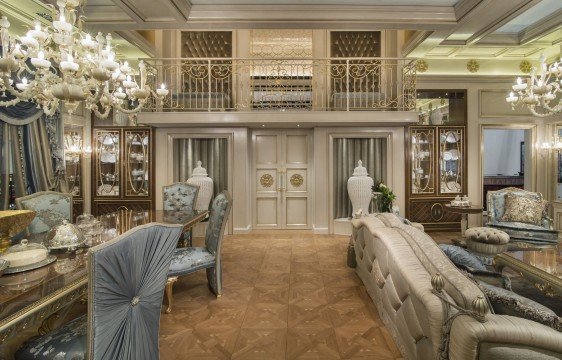 This picture shows a modern, white and marble kitchen. The kitchen features an open shelving design with marble countertops, stainless steel appliances, white cabinetry, and a large island with marble top. An archway leads to another area of the kitchen, which appears to have an eat-in dining area. The room also features tall glass windows that allow natural light to fill the space.