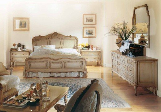 This picture shows a large, bright open space with a wall of windows letting in plenty of light. There is a white sofa against one wall and wooden furniture with bookshelves filled with books and art. The floor is covered with a patterned area rug, and there are several plants spaced throughout the room.