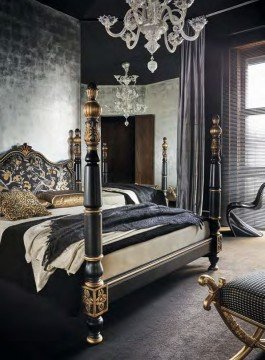 This picture shows a stunning and luxurious master bedroom with a grand regal aesthetic. The room has walls upholstered with a red velvet fabric with gold gilded detailing, and opulent draperies of the same fabric. The floor is covered in a deep brown wood parquet and features a traditional red and gold Aubusson area rug. The center of the room is dominated by a large bed upholstered with a white and gold patterned fabric, and matching throw pillows. On the left side of the bed is an intricately carved wooden nightstand, and on