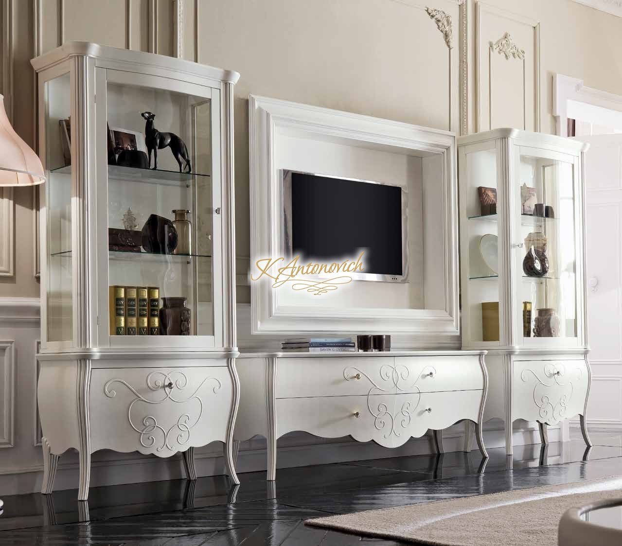 This picture shows a modern, luxury living room with white walls and a black and gold checkered marble floor. The living room features two black velvet sofas arranged facing each other, with a large gold-rimmed glass coffee table between them. The room also includes an ornate gold and crystal chandelier suspended from the ceiling, and a large gold and white framed mirror mounted on the wall. On the coffee table are several books, and there is a black and white patterned area rug beneath the furniture.