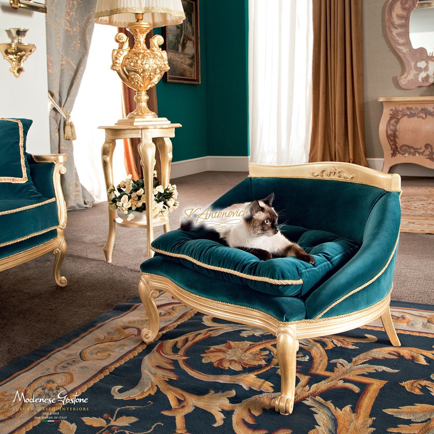 This picture shows a modern, luxury living room. The walls are painted white, and the floor is a contrasting dark hardwood. There is a large, comfortable-looking grey upholstered sofa with several colourful and patterned throw pillows arranged around it. On either side of the sofa are tall black and gold lamps, and behind it is a minimalistic wall display with black frames. In the corner is an oval-shaped blue velvet ottoman and in front of that is a glass end table with a white vase filled with bright yellow flowers.