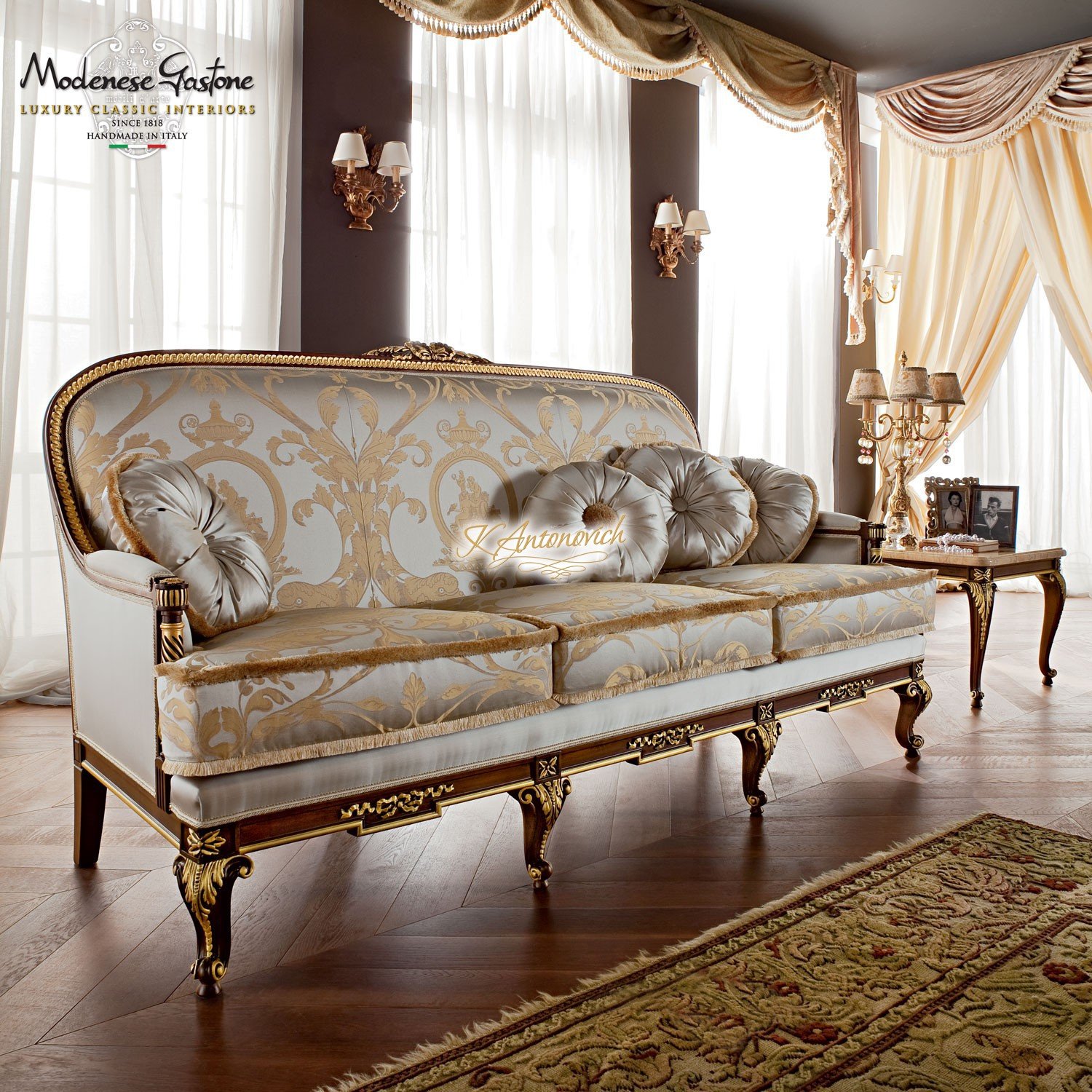 This picture shows a beautifully designed interior for a luxury living room. The colors used in the design include warm earth tones such as beige and brown, as well as gold accents. The room features an elegant sofa set with ornate armrests, one of which has a decorative side table next to it. On the opposite wall is a large mirror framed in gold and an artwork hung in between. The floor is covered with a plush cream-colored carpet, while the ceiling is lit by a unique chandelier suspended from an intricate network of metal beams.