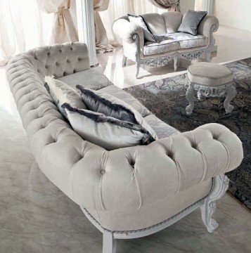 This picture shows a modern and luxurious living room designed by Antonovich Design, an interior design company based in the United Arab Emirates. The room features a plush velvet couch with accent pillows, a large ornate chandelier, and a stunning marble-tiled floor. There is also a unique marble wall with inlaid gold trim, a rustic area rug, and a built-in entertainment center, featuring a large flat-screen TV and shelving for storage. The overall look of the room creates an elegant and inviting atmosphere.