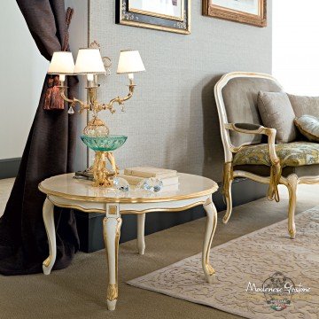 This picture is of an ornate, opulent bedroom designed in a modern-minimalist style. The room features a four-poster bed with a white teal and pink patterned headboard and sides, surrounded by sheer curtains. A grey sofa is placed in front of the bed and there is a tall lamp with a glass base next to it. On the other side of the bed is a large crystal chandelier, and there are grey and gold armchairs placed against the walls. The walls are white and there is a rug with a geometric pattern on the floor.