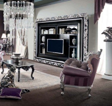 This picture shows a luxurious living room with a light gray velvet couch and armchairs in the center. The walls are light beige decorated with gold colored wall frames, and a crystal chandelier hangs from the ceiling. On the left side is a wooden stand with a decorative plant, and there are two modern black marble tables on either side of the seating area. The window is draped in heavy white curtains, and the floors are tiled with a geometric pattern.
