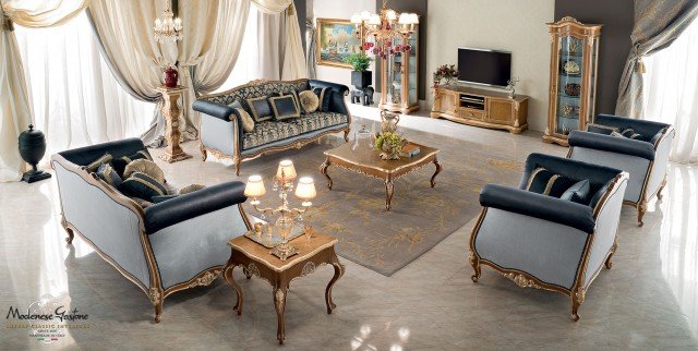 This picture shows an elegant living room with a luxurious and modern design. The room has a light beige color scheme and is decorated with glossy white furniture, a modern fireplace, and crystal chandeliers. The floor is covered in a cream-colored carpet, while the walls display beautiful artwork. A few comfortable armchairs, ottomans, and other accents provide a cozy atmosphere.