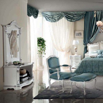 The picture shows a luxurious bedroom with an impressive aesthetic. The room features a four-post bed with a white and beige bedspread topped with a variety of accent pillows. The room is decorated with a mirrored wall, marble flooring, a grey tufted chaise lounge chair, and two chairs upholstered in a matching beige fabric. Several lamps are scattered about the room to provide different levels of lighting and accent pieces, including an elegant chandelier.