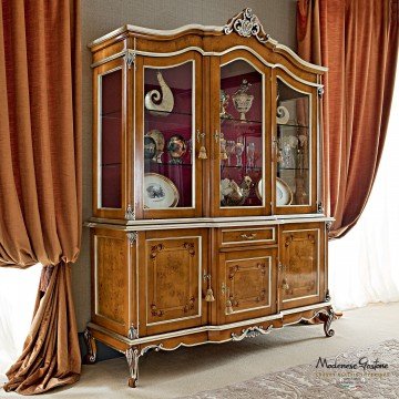 This picture shows a luxurious neoclassical living room with stunning gold and white decor. The room features an intricately carved cream-colored sofa that sits atop a white rug, while two large windows are adorned with elaborate gold curtains. The walls are painted a rich shade of blue and feature several classical oil paintings. A grand chandelier hangs from the ceiling, providing bright lighting and a regal atmosphere.