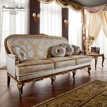 This picture shows a beautifully designed interior for a luxury living room. The colors used in the design include warm earth tones such as beige and brown, as well as gold accents. The room features an elegant sofa set with ornate armrests, one of which has a decorative side table next to it. On the opposite wall is a large mirror framed in gold and an artwork hung in between. The floor is covered with a plush cream-colored carpet, while the ceiling is lit by a unique chandelier suspended from an intricate network of metal beams.