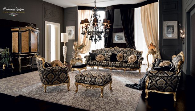 This picture shows an elegant and modern dining room, featuring a black and white color palette. The walls are painted in a dark grey, with white trim and crown molding. There is a sleek, white dining table with black chairs, illuminated by an ornate, crystal chandelier. The furniture is finished in gold and the room is accented with a flower vase, mirrored tray, and a gilded framed art piece.