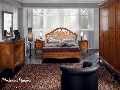 This picture shows a luxurious bedroom with a four-poster bed. The bed has several plush, patterned pillows and a matching headboard that is upholstered in a deep navy fabric. The bedding is made of a rich, dark velvet, and there are several lighter throw pillows with geometric designs. The walls are a light, muted blue color with intricate wood paneling. There are two end tables on either side of the bed, with tall golden lamps and accent pieces on top. The room has a large area rug with a patterned print, and two sheer