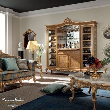 This picture shows a luxurious bedroom in an interior design style called Baroque. The walls and floor are a light cream color and the furniture has an ornate and lavish look to it, featuring gold accents and patterns. The bed is upholstered in a white and gold fabric and is paired with nightstands featuring intricate carved details. In the corner of the room is an elegant lounge chair with an ottoman and a chandelier hangs from the ceiling. There is a large mirror above the bed and a set of French doors leading out to what appears to be a balcony