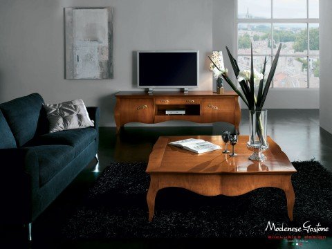 The picture shows a large rustic wooden table surrounded by four chairs in a cozy and warm room. The walls are painted a deep red color, and there is a fireplace with a white mantle, accented by several potted plants. The cushioned chairs are upholstered in a pattern of red, cream, and brown. On the table is a vase filled with fresh flowers, adding a touch of brightness to the room.