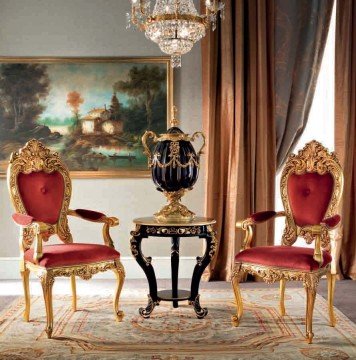 This picture shows a luxurious living room with a cream-colored couch and armchair in the center. The walls are painted a deep red color with white crown molding along the top. The floor is made of light-colored marble and there is a glass coffee table with a black vase and flowers in the center. On the left side of the room is a large window framed with ornate white curtains. A potted plant sits on the window sill and there is an ornately carved mirror on the right wall.