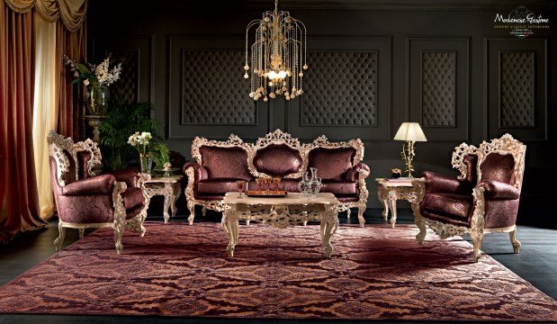 This picture shows a luxurious living room with a black and gold color scheme. There is a large tufted couch with multiple black and gold cushions, a white coffee table with a shiny gold frame, a large mirror on the wall with intricately carved gold details, and two black chairs with gold legs. The walls are a light grey with a beige area rug to complete the look.