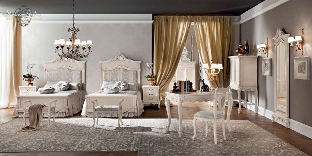 This picture shows a luxurious bedroom designed in an opulent style. The room features lavish touches such as a custom four-poster bed with light-colored velvet bedding, ornately framed paintings, mirrored furniture and an intricately woven rug. A glass chandelier hangs from the ceiling, illuminating the room with its warm, inviting glow. The walls are painted a pale shade of pink, adding to the overall soft and elegant feel of the space.