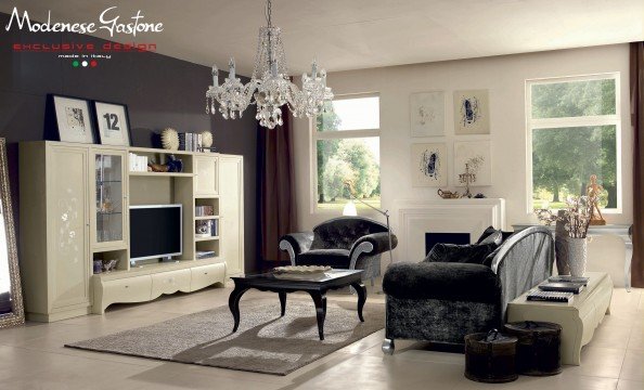 This picture is of a modern home interior featuring a sophisticated living room with white walls and gray furniture. The room features a black velvet sofa, a white marble coffee table, a snakeskin ottoman, and a large flat-screen TV mounted above the fireplace. There is also a metal and glass ceiling fan in the center of the room and two art pieces hanging on the walls. The room is accented with patterned pillows, mirrored side tables, and an antique floor lamp.