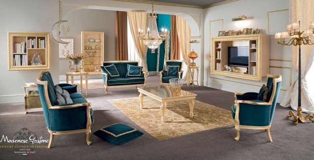 This picture shows a luxurious modern living room with a grey and white color scheme. The seating area features a large sectional sofa with a chaise lounge, two arm chairs, and an over-sized ottoman. The walls are adorned with contemporary art pieces and the room is illuminated with an overhead chandelier and sconces. A glossy black coffee table sits in the middle of the room, and there is also a patterned rug for added warmth.