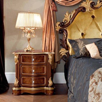 This picture shows a luxurious master bedroom with a four-poster, tufted bed with a gilded headboard. The bedding is a combination of blue velvet and silk bedding with a grey furry blanket. There is an armchair and table to one side with a lamp, as well as a floor-to-ceiling padded window seat with a view of the ocean beyond. The walls are mostly white with a subtle patterned wallpaper in silver and off-white, supplemented by wall art and recessed lighting. The room is decorated with potted plants and soft, warm