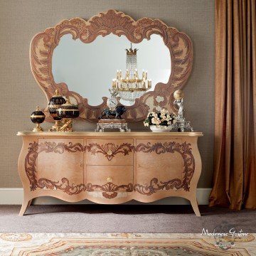 This picture shows a large, white and gold bedroom with a luxurious design. There are ornate white curtains, mirrors on the wall, a floral patterned wallpaper, a white and gold upholstered bed with a white tufted headboard, a white leather chair with gold legs, and a glass-top side table with gold accents. To one side of the room is a large window allowing plenty of natural light to come in.
