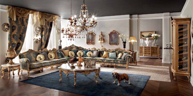 This picture is of an interior design featuring a modern, luxurious living room. The room is accented with gold accents and includes a plush sofa, two armchairs, coffee tables, and end tables. It also includes framed art hanging on the wall, as well as tall floor lamps with crystal details that add a sparkle to the space. The colors in the room are largely neutral, including white and beige, along with metallic accent colors of gold, silver, and bronze giving the room a glamorous feel.
