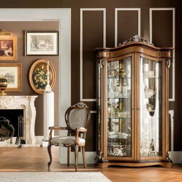 This picture shows a grand two-story entry foyer with an elegant glass spiral staircase. The walls are adorned with large mirrors and wall sconces, and the floor is covered in a light-colored marble. A large, teardrop-shaped chandelier hangs from the ceiling, adding to the room's luxurious atmosphere.