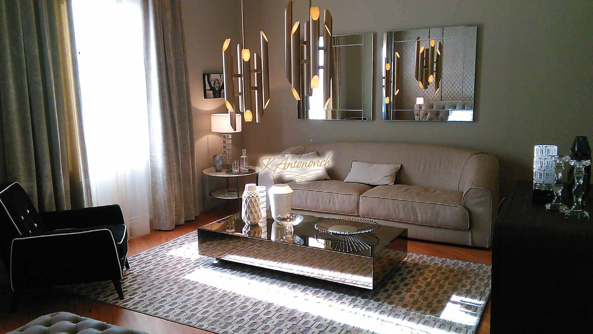 This picture shows a luxurious, modern living room designed with ornate decorations and high-end furnishings. The space is decorated with a light coloured palette with hints of gold in the furniture, and contrasting brown and beige hues in the walls and floor. A large chandelier hangs from the ceiling, creating an opulent effect. An upholstered sofa and armchair set provides a comfortable seating area, while additional seating is provided by two small stools near the window. A glass coffee table, side tables, and various decorative pieces such as lamps and vases complete the