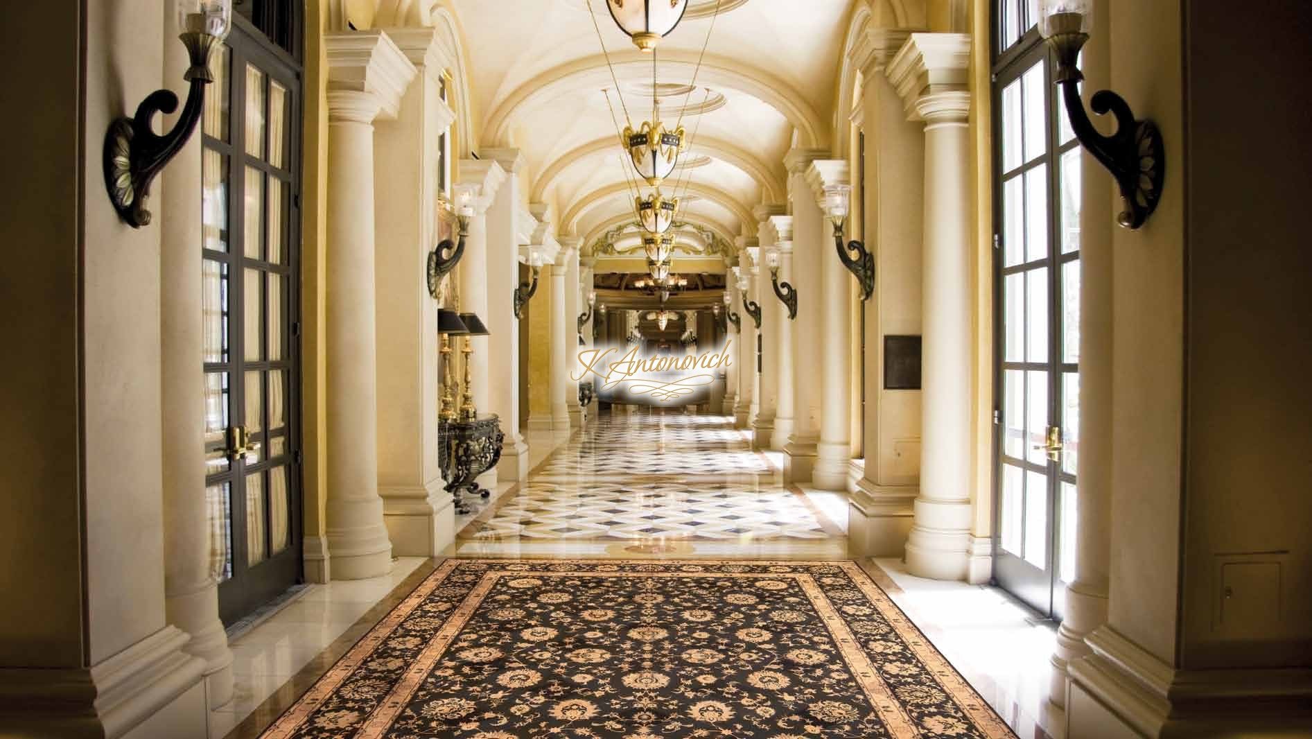 This picture shows an elegantly designed interior hall with a grand staircase. The walls are painted in a neutral beige color and have intricate white molding around the edges. The stairs feature two white marble railings on either side, as well as a detailed wrought-iron balustrade. The stairs themselves are wooden and feature intricately carved patterns along their length. A large chandelier hangs from the ceiling and provides ambient lighting to the room. The area is also decorated with mirrors, statues, and vases, giving it a luxurious atmosphere.