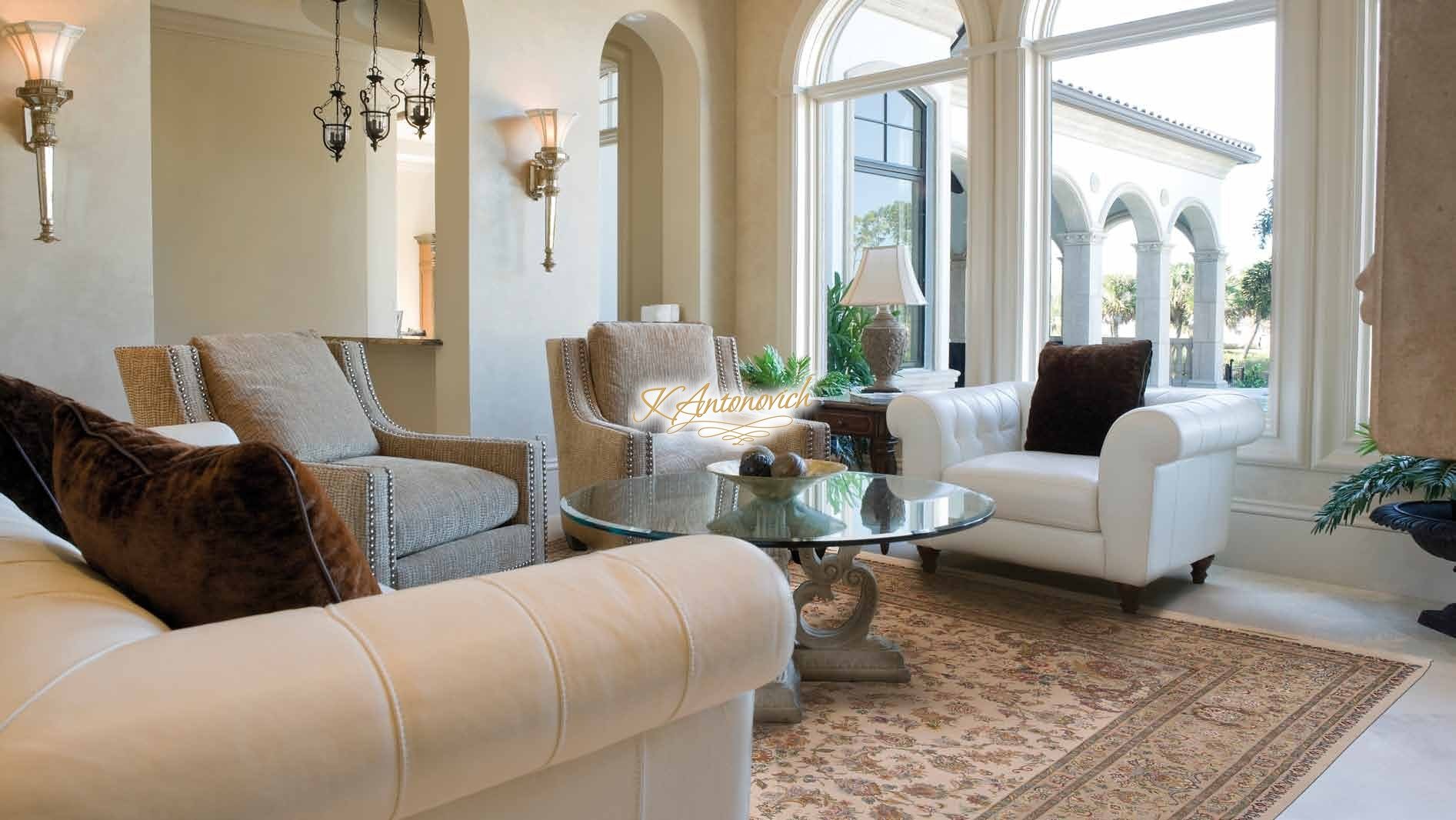 This picture shows a luxurious living room decorated with gold and white accents. The room features a large ornate couch, two white armchairs, a glass coffee table, and a white shag rug. The walls are adorned with gold-framed mirrors, and the ceiling has recessed lighting. The overall look is elegant and sophisticated.