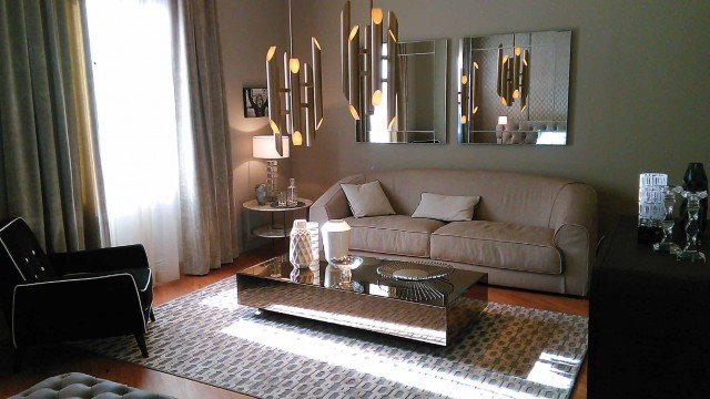 This picture shows a luxurious, modern living room designed with ornate decorations and high-end furnishings. The space is decorated with a light coloured palette with hints of gold in the furniture, and contrasting brown and beige hues in the walls and floor. A large chandelier hangs from the ceiling, creating an opulent effect. An upholstered sofa and armchair set provides a comfortable seating area, while additional seating is provided by two small stools near the window. A glass coffee table, side tables, and various decorative pieces such as lamps and vases complete the