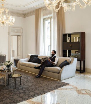 This picture shows a luxurious, open-concept living area with marble floors and modern furnishings. There is a dark wood dining table with white armchairs, a white sofa and two patterned armchairs, and a marble-top sideboard. The walls are covered in white wallpaper with a neutral striped pattern. On the wall above the sofa, there is a large gold mirror and on either side, tall black lanterns hang from the ceiling. The room is filled with natural light from the large windows on the left-hand wall.