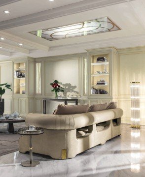 This picture shows a white, modern living room with luxurious furniture. There is a white sofa in the center of the room and two matching white armchairs on either side of it. The walls and ceiling are painted white and accented with a silver chandelier. There is also a glass coffee table, a white rug, and several plants throughout the room.